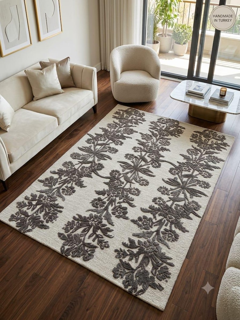 May include: A rectangular area rug featuring a cream-coloured base with three vertical columns of dark grey, raised floral and leaf patterns. The rug has a textured pile and includes a circular tag in the corner that reads Handmade in Turkey.