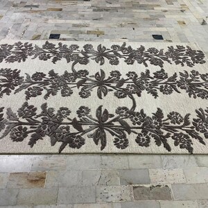 May include: A rectangular area rug featuring a cream-coloured base with three horizontal bands of dark brown floral patterns. The design consists of symmetrical, repeating botanical motifs including leaves and blossoms arranged in a linear fashion.