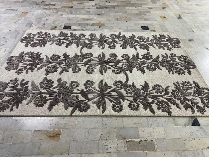 May include: A rectangular area rug featuring a cream-coloured base with three horizontal bands of dark brown floral patterns. The design consists of symmetrical, repeating botanical motifs including leaves and blossoms arranged in a linear fashion.