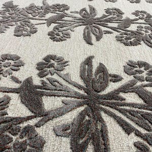 May include: A textured area rug featuring a light beige background with a raised, dark brown floral pattern. The design consists of interconnected flowers, leaves, and stems that create a dense, repeating botanical motif across the surface.