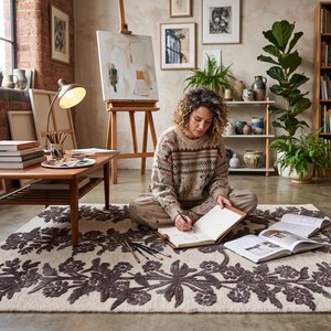 May include: A rectangular area rug featuring a cream-coloured base with a prominent, dark charcoal-grey floral and vine pattern embroidered across the surface. The rug has a textured, tufted appearance with a repeating botanical motif.
