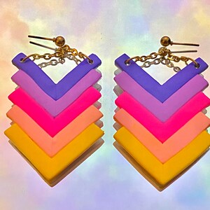 May include: A pair of colourful chevron earrings with a gold chain and hook. The earrings are made of polymer clay and feature a gradient of yellow, orange, pink, purple, and lavender.