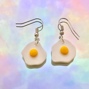 May include: A pair of silver dangle earrings with white and yellow fried egg charms.