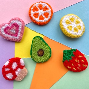May include: Six felt food patches in various shapes and colours. A pink heart, a green avocado, a red and white mushroom, a yellow lemon, an orange orange, and a red strawberry.