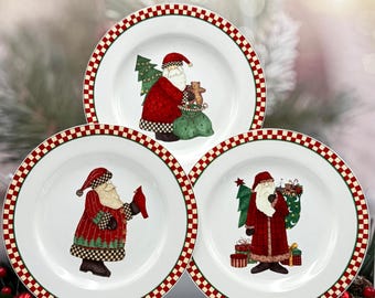 Puede incluir: Tres platos de cerámica blancos con un borde a cuadros rojo y verde. Cada plato presenta una ilustración diferente de Papá Noel con un abrigo rojo, sosteniendo regalos y un árbol de Navidad. Decoración festiva navideña.