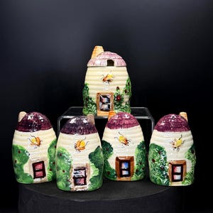 May include: A collection of ceramic beehive-shaped containers. The containers are painted with a cream-colored base, green foliage, and a purple roof. Each has a small window and a painted bee. One container has a lid.