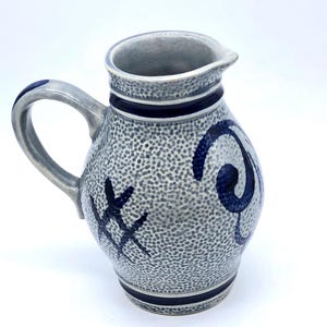 May include: Grey ceramic jug with a handle and spout. The jug has a textured surface and is decorated with dark blue bands and abstract designs. The jug is a decorative item.