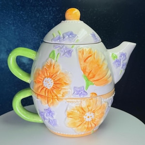 Puede incluir: Juego de té de cerámica blanca con tetera y taza. Decorado con diseños florales naranjas y morados, con un asa verde. La tetera tiene una tapa con un pomo naranja. La taza está apilada debajo de la tetera.