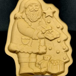 Op de afbeelding: Tan-kleurige kerstman koekjesvorm met een ingepakt cadeau en naast een kerstboom met een ster erboven. Een teddybeer en een ander cadeau staan aan de voeten van de kerstman. De vorm heeft een gat aan de bovenkant om op te hangen.