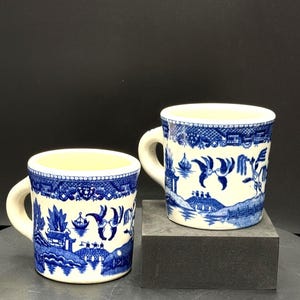 Op de afbeelding: Twee keramische mokken met een blauw-wit Willow-patroon. De mokken hebben een crèmekleurige binnenkant en een gebogen handvat. Het ontwerp toont een landschap met vogels, bruggen en gebouwen. De mokken staan op een donkergrijze ondergrond.