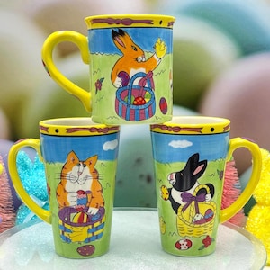 May include: Three ceramic Easter mugs with yellow handles and rims. Each mug features a different cartoon animal: a rabbit, a cat, and a bunny, all holding Easter baskets. The mugs have a green grass background and a blue sky.