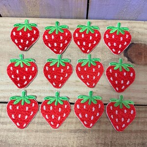 May include: Twelve embroidered strawberry patches. Each patch features a red strawberry with white seeds and a green stem and leaves. The patches are arranged on a light brown wooden surface.