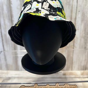 May include: A black bucket hat with a white, black, yellow, and teal controller pattern. The hat is displayed on a black mannequin head. The brim is black and the hat is reversible.