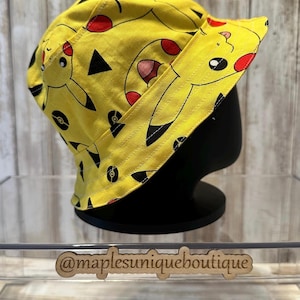 May include: Yellow bucket hat featuring a Pikachu pattern with black outlines and red accents. The hat is displayed on a black mannequin head. The text "@maplesuniqueboutique" is visible on the display.