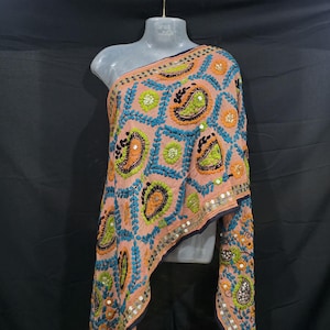 May include: A peach shawl with detailed embroidery. It features paisley and floral patterns in blue, green, and orange, with small mirrors. The shawl is draped over a mannequin, showcasing its design.