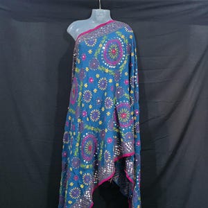 May include: A blue shawl with detailed embroidery and mirror embellishments. The shawl displays a repeating pattern of circular designs in purple, pink, and yellow, with small star accents. The edges are finished with a pink border.