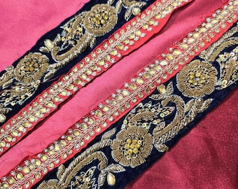 May include: Close-up of two decorative fabric strips. One strip is navy blue with gold floral embroidery and sequins. The other strip is red with gold embellishments. The background is a pink fabric.