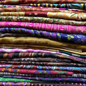 May include: A stack of colorful fabric scraps with various patterns and designs. The fabrics are folded neatly and arranged in a vertical stack. The colors include shades of purple, pink, brown, green, and gold.