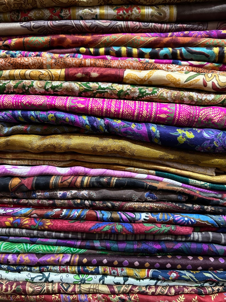 May include: A stack of colorful fabric scraps with various patterns and designs. The fabrics are folded neatly and arranged in a vertical stack. The colors include shades of purple, pink, brown, green, and gold.