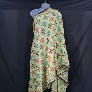 Könnte beinhalten: Ein hellgrüner Schal mit einem Blumenmuster in Rot, Orange, Blau und Braun. Der Schal hat einen schwarzen Rand mit Spiegelverzierungen. Der Stoff ist über eine Schaufensterpuppe drapiert, um das Muster und die Textur hervorzuheben.