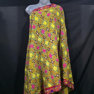 Könnte beinhalten: Ein drapiertes Textil mit einem Blumenmuster in Gelb, Rosa und Blau auf grünem Hintergrund. Der Stoff hat einen roten Rand mit aufwendigen Details. Das Textil wird auf einer Schaufensterpuppe präsentiert.