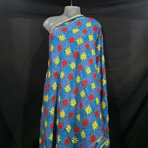 Könnte beinhalten: Blauer Stoff mit einem Blumenmuster aus roten und gelben Blüten. Der Stoff ist über eine Schaufensterpuppe drapiert. Die Ränder sind gelb eingefasst. Der Hintergrund ist schwarz.