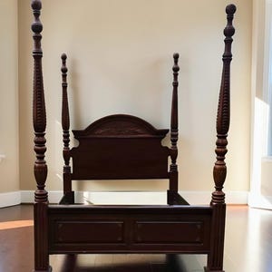 May include: A dark brown wooden four-poster bed frame featuring tall, intricately carved posts with rounded finials. The headboard has a curved top edge with subtle floral carvings, while the footboard displays two rectangular recessed panels.