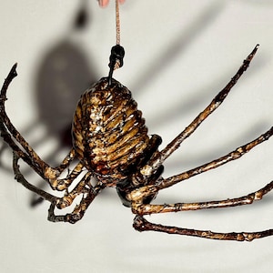 Op de afbeelding: Een bruine en zwarte spin met lange poten die aan een touwtje hangt. De spin is gemaakt van papier-maché en heeft een gedetailleerd, getextureerd oppervlak.