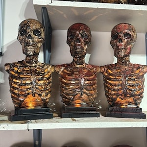 May include: Three skeletal figures with exposed ribs and a fleshy exterior, each with a different facial expression. The figures are displayed on a shelf with other similar figures.