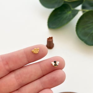 May include: Two small bee figurines, one is yellow and the other is white with black stripes. The figurines are on a person's hand.