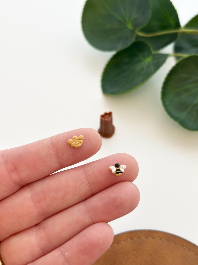 May include: Two small bee figurines, one is yellow and the other is white with black stripes. The figurines are on a person's hand.