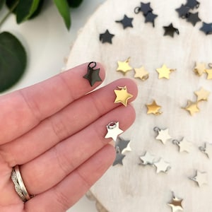 Op de afbeelding: Een verzameling stervormige bedels in verschillende metallic afwerkingen: zwart, goud en zilver. De bedels zijn klein, met een lus aan de bovenkant om aan sieraden of andere knutselwerken te bevestigen. Ze worden gepresenteerd op een lichtgekleurd houten oppervlak.