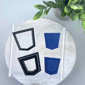 May include: Two clear acrylic sheets with two black plastic stencils and two blue felt shapes. The stencils are shaped like a pocket with a rounded top. The felt shapes are the same shape as the stencils.