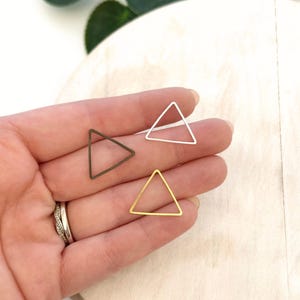 May include: Three geometric triangle charms in bronze, silver, and gold tones. The charms are held in a hand, showcasing their delicate, open-frame design. The background is a soft, neutral color, highlighting the jewelry.