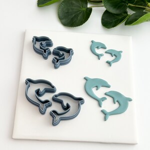 May include: A set of four dolphin cookie cutters, two are gray and two are light blue. The cutters are on a white surface.