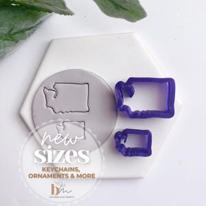 May include: Two purple plastic cookie cutters in the shape of the state of Washington. The cutters are on a white hexagonal surface. The text "new sizes" is written in black on a white circle. The text "keychains, ornaments & more" is written in black below the circle. The text "bm" is written in black on a white rectangle below the text.