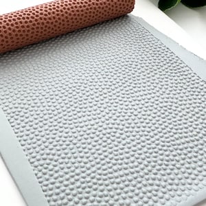 May include: A terracotta-colored textured roller rests on a gray, dotted surface. The roller has a pattern of small, evenly spaced holes. The gray surface is also covered in a similar pattern of small, raised dots, creating a tactile effect.