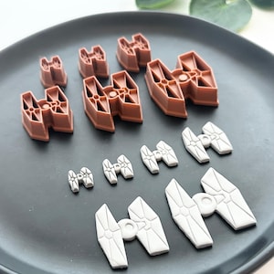 May include: A collection of brown and gray TIE fighter-shaped decorations on a black plate. The decorations vary in size, with the brown ones larger than the gray ones. The shapes are arranged in rows, showcasing a space-themed design.