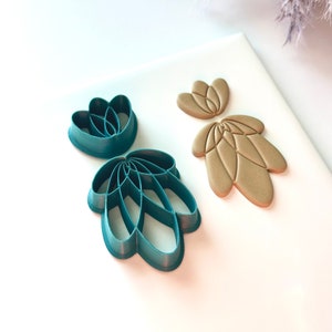 Pode incluir: Três cortadores de argila polimérica verde-azulado em forma de flores, ao lado de duas peças de argila bege. Os cortadores têm a forma de flores e folhas estilizadas. As peças de argila têm a forma de uma flor e uma folha.