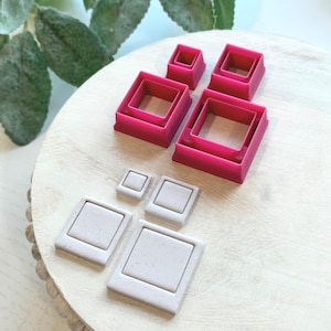 Pode incluir: Conjunto de oito cortadores de biscoitos quadrados de plástico rosa e branco. Os cortadores estão dispostos em círculo sobre uma superfície de madeira branca.