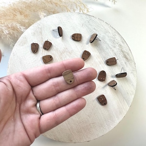 May include: Wooden stud earrings in a variety of shapes, including rectangles and rounded rectangles. The earrings are a warm brown color and are displayed on a white, textured surface. Some earrings are shown with the earring posts attached.