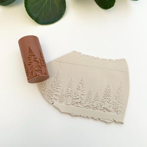 May include: A brown rolling pin with a raised design of a forest with trees. The rolling pin is used to create an impression in clay.