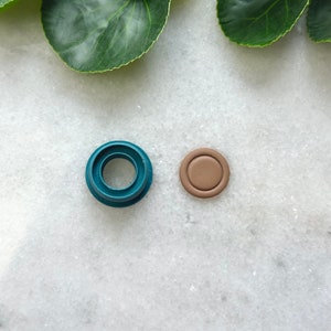 May include: A teal blue plastic ring mold and a brown clay circle, both on a white marble surface.