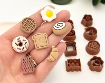 Könnte beinhalten: Ein Set mit 10 braunen und weißen Ausstechern in Form von Frühstücksgerichten, darunter ein Donut, eine Waffel, ein Croissant, ein Zimtschnecken, ein Spiegelei, eine Scheibe Brot und Speck.