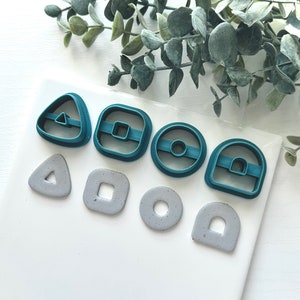 May include: A collection of teal-colored polymer clay cutters in various geometric shapes, including a triangle, square, circle, and arch. Below are matching gray clay pieces. The cutters and clay are arranged on a white surface with green foliage.
