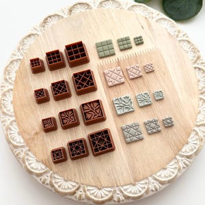 May include: A collection of square-shaped clay cutters in various sizes and designs. The cutters are brown, green, and pink, featuring grid, geometric, and floral patterns. They are arranged on a light wooden tray with a decorative edge.