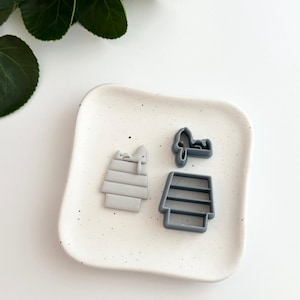 May include: Three gray silicone cookie cutters shaped like a beehive, a bee, and a honeycomb. The cutters are on a white speckled plate.