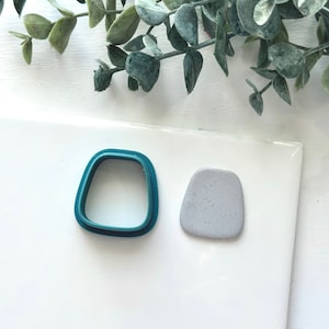 May include: A teal plastic cookie cutter shaped like a rounded rectangle next to a gray clay shape that has been cut out with the cookie cutter.