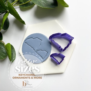 Op de afbeelding: Twee paarse plastic koekjesvormen in de vorm van de staat South Carolina. De vormen liggen op een witte zeshoekige tegel met een blauwe kleiafdruk van de staat South Carolina. De tekst "new sizes" en "keychains, ornaments & more" is op de tegel gedrukt.