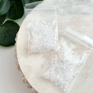 Pode incluir: Duas sacolas de plástico transparentes cheias de pequenas peças de plástico transparente para a parte de trás dos brincos. As sacolas estão sobre uma superfície de madeira branca.